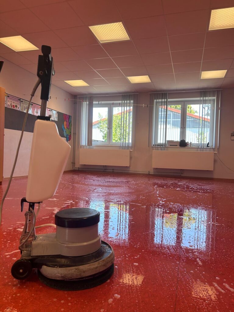 Einscheibenmaschine auf rotem Boden in einem Gruppenraum einer Kita während der Grundreinigung, Schaum und Feuchtigkeit auf der Bodenfläche sichtbar, im Hintergrund große Fensterfront.