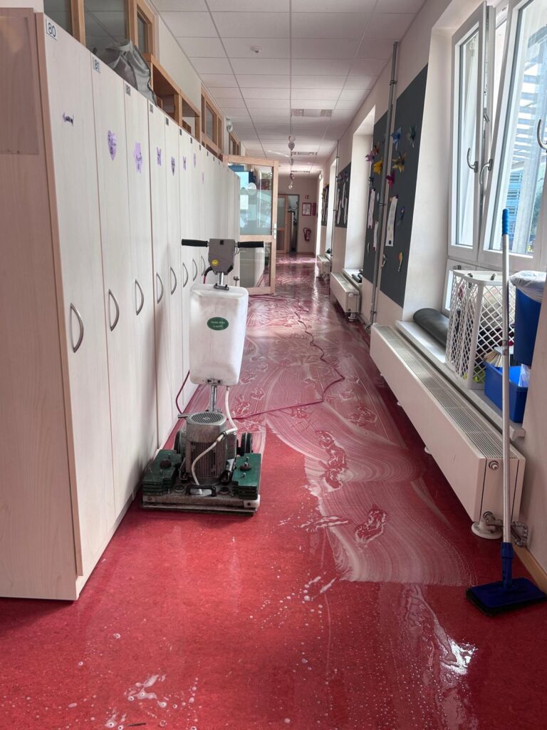 Maschine zur Grundreinigung auf einem roten Bodenbelag in einem Kita-Flur, umgeben von Garderobenschränken, offenem Fenster und Reinigungsequipment.