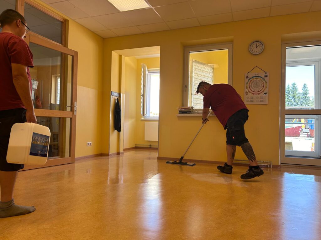 Ein Mitarbeiter trägt in einem Kita-Raum mit Flachmopp eine Pflegeemulsion auf den frisch gereinigten Linoleumboden auf, während ein Kollege die Kanister mit dem Bodenpflegemittel hält.