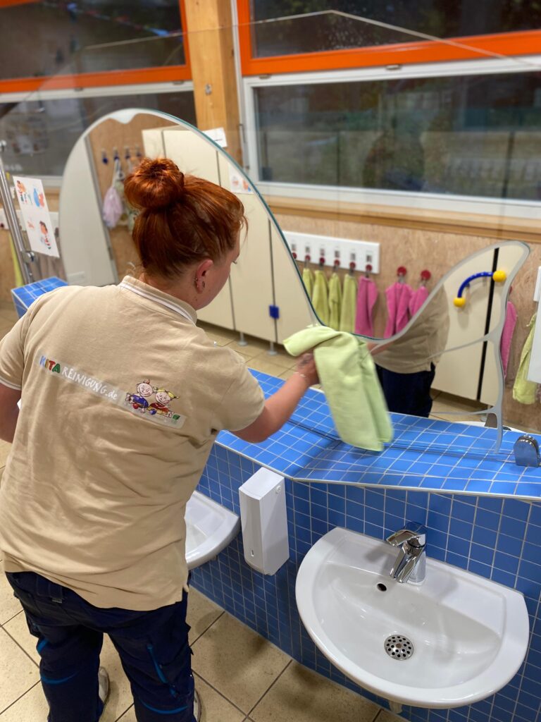 Reinigungskraft von Kitareinigung.de beim Putzen eines Spiegels im Waschraum einer Kita mit kindgerechter Ausstattung, blau geflieste Waschplätze und Haken für Handtücher im Hintergrund.