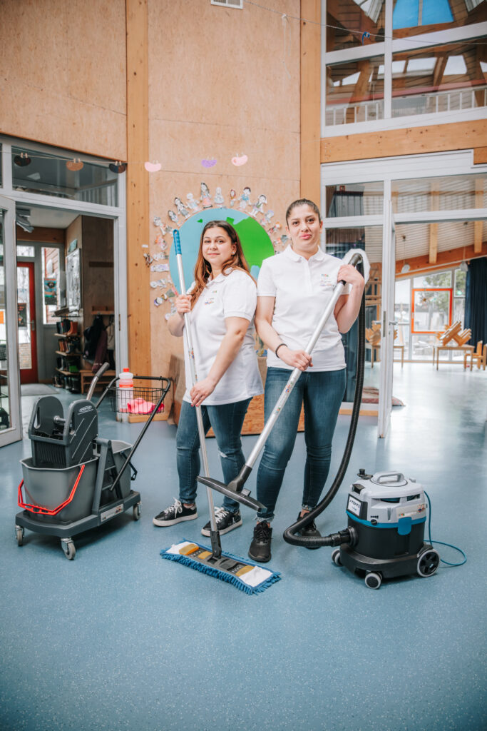 Zwei Mitarbeiterinnen in Arbeitskleidung mit Reinigungsgeräten in einer modernen Kita, bereit zur Unterhaltsreinigung