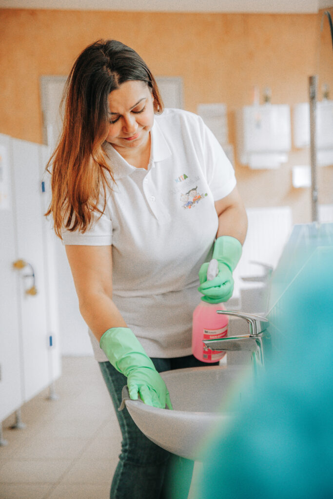 Reinigungskraft mit grünem Handschuh und Desinfektionsmittel reinigt ein Waschbecken im Sanitärbereich einer Kita, trägt dabei ein weißes Polo mit Logo von Kitareinigung.de.