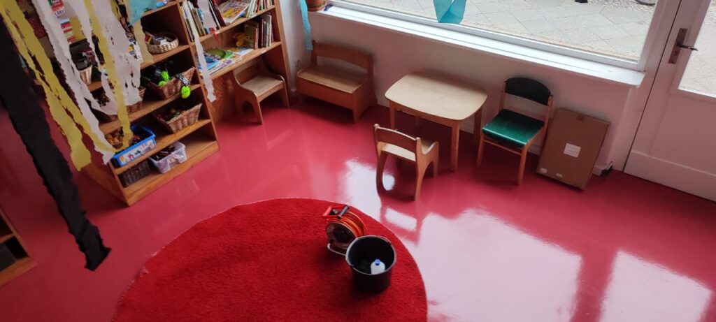 Spielbereich in einer Kindertagesstätte mit frisch gepflegtem rotem Bodenbelag, Kindermöbeln und Teppich – im Vordergrund Eimer und Schlauch als Reinigungsutensilien.